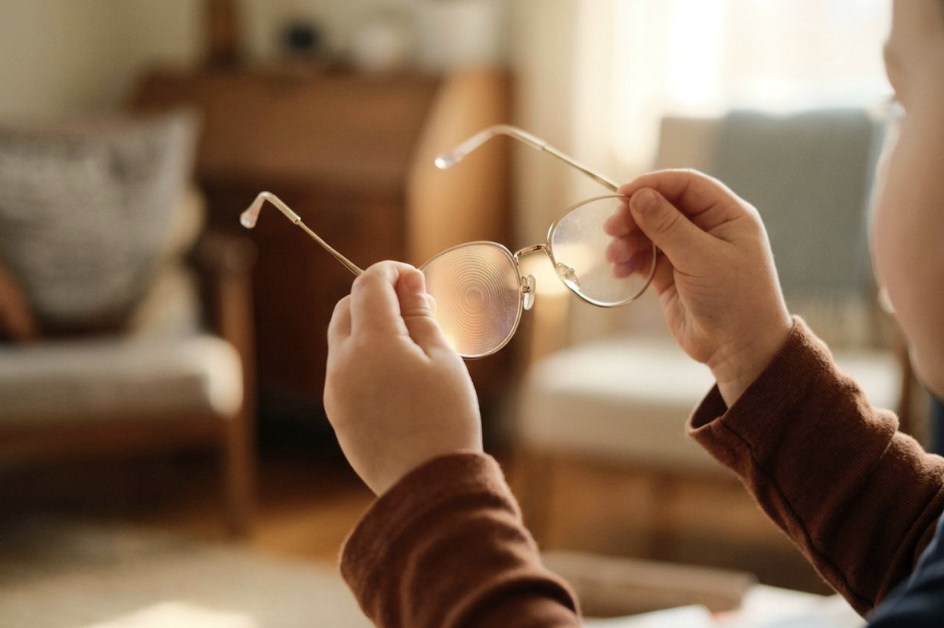 A child holds wire-rimmed eyeglasses indoors. Light reflects off one lens, revealing a distinct pattern of concentric circles.